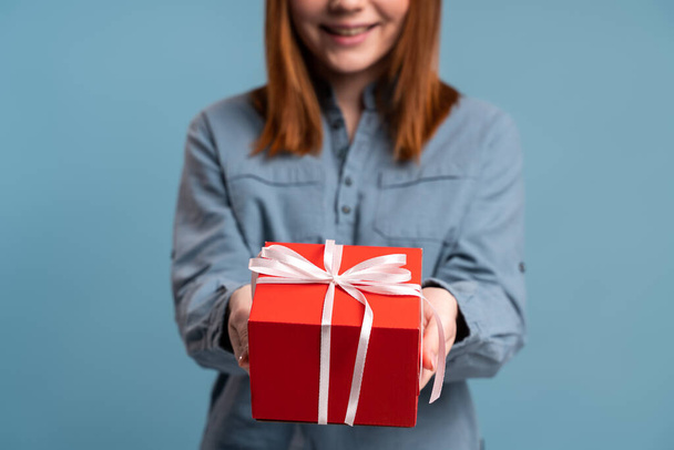 笑顔の女性は、プレゼントを与え、現在のボックスを保持、楽観的な表現でカメラを見て、挨拶。青い背景に隔離された屋内スタジオショット  - 写真・画像