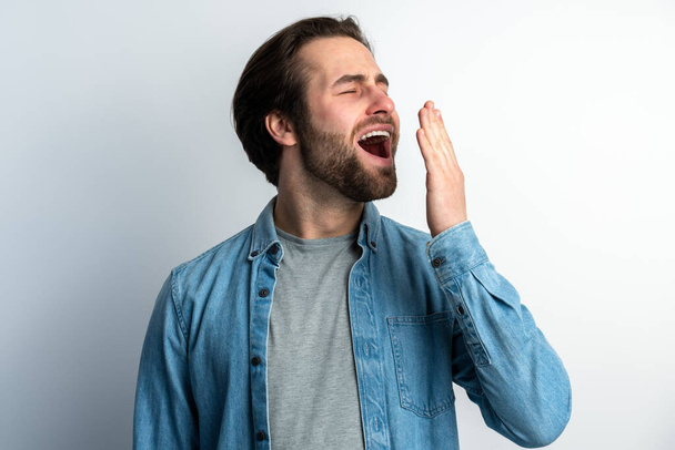 I need rest. Portrait of sleepy brunette man with beard yawning and covering mouth with hand, feeling exhausted, lack of sleep. Indoor studio shot isolated on white background  - Fotoğraf, Görsel