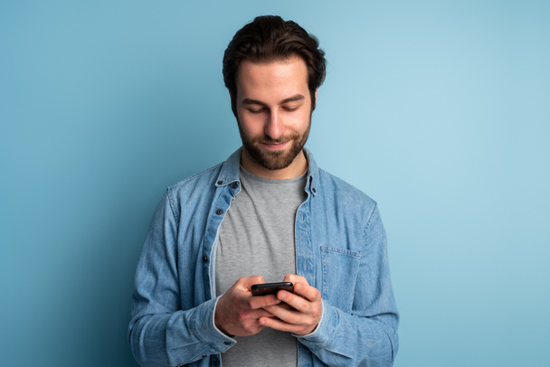 Portrait of cheerful brunette man with beard using cellphone and smiling, reading good news message, enjoying mobile application. Indoor studio shot isolated on blue background  - Photo, image