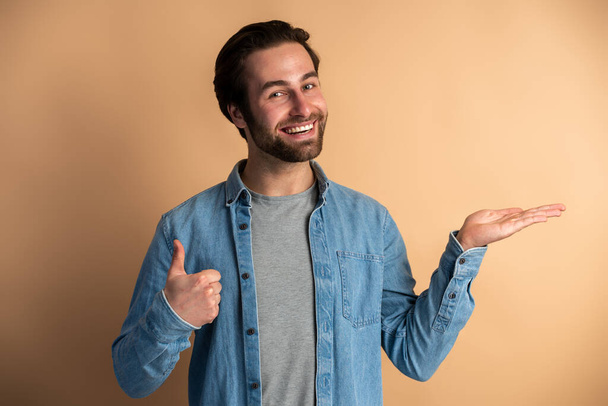 Handsome man presenting copy space on his palm and looking happy, showing empty place for promotional text. Indoor studio shot isolated on orange background  - Foto, Bild