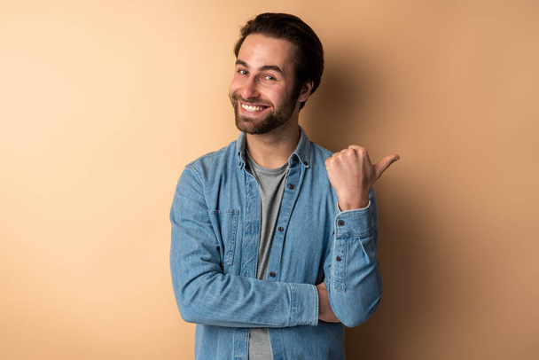 Advertise here. Positive bearded man pointing finger away and paying attention at empty space for advertisement, while looking at camera with toothy smile. Indoor studio shot isolated on orange background  - Foto, Imagem