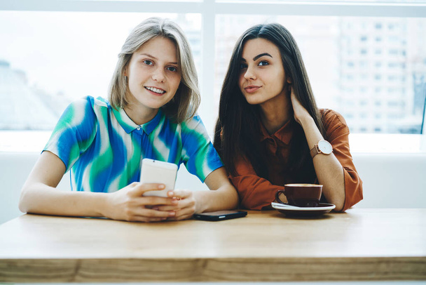 Positive female friends in casual clothes sitting at table with smartphone and cup of coffee looking at camera spending time together - Foto, immagini