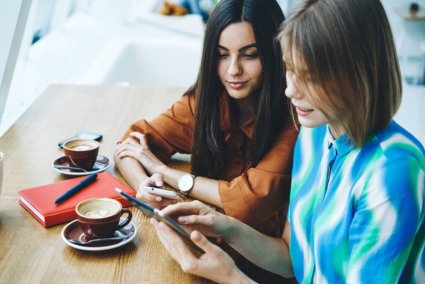High angle of young female friends using tablet together while sitting in modern cafe at table with cups of coffee and notebook and with smartphone in hand - Фото, зображення