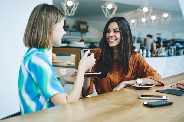 Young multiethnic female friends laughing and discussing news looking at each other while having hot beverage sitting at wooden table with gadgets in modern cafeteria - Foto, Imagem