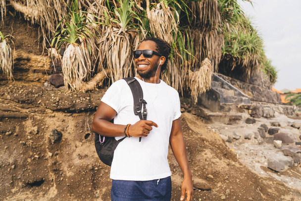 Happy African American male in casual clothes and backpack standing near cliff and looking away while listening to music in earphones - Фото, зображення