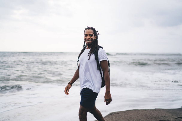 Joyful African American male in casual outfit with backpack smiling and looking at camera while standing on seashore in summer - Foto, imagen