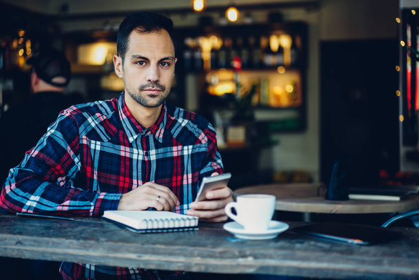 Half length portrait of trendy dressed customer with planner textbook and modern smartphone device for checking web news looking at camera during weekend time in cafe interior, handsome blogger posing - Foto, Imagen