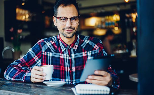 Portrait of handsome man looking at camera during coffee break for updating application on digital tablet, Caucasian male blogger in eyewear connecting to free 4G high speed internet on gadget - Fotó, kép