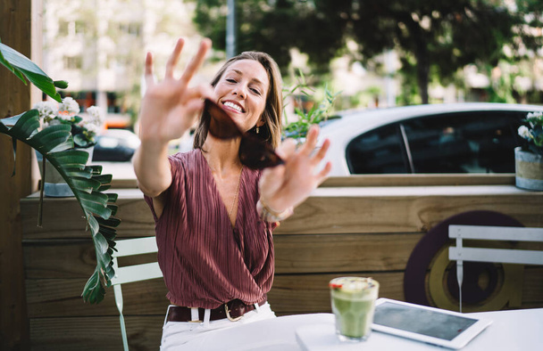 Happy caucasian female in trendy wear sitting on cafe terrace holding sunglasses enjoying free time, smiling woman satisfied with free time on terrace with green smoothie recreating during work break - Фото, зображення