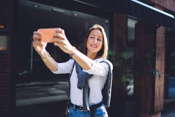 Cheerful 40 years old lady in trendy outfit smiling and taking self portrait on smartphone while standing on street on sunny day - Photo, image