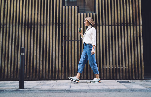 Side view of stylish positive middle aged female in jeans and sneakers strolling along street near modern building and drinking coffee - Foto, immagini