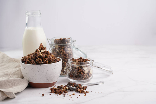 Breakfast muesli cereal with chocolate chips and nuts in several jars and bowl, bottle of milk, white surface - 写真・画像