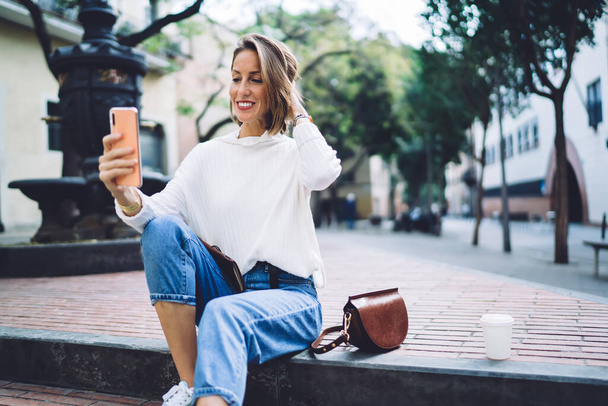 Positive female tourist in trendy outfit taking self portrait on mobile phone while relaxing on stairs of park against fountain after sightseeing - 写真・画像