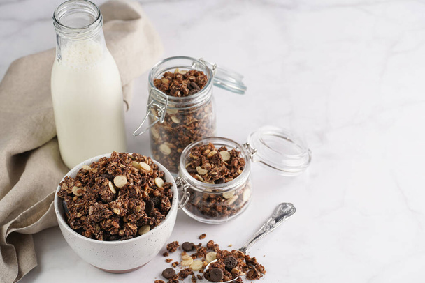 Breakfast muesli cereal with chocolate chips and nuts in several jars and bowl, bottle of milk, white surface - Zdjęcie, obraz