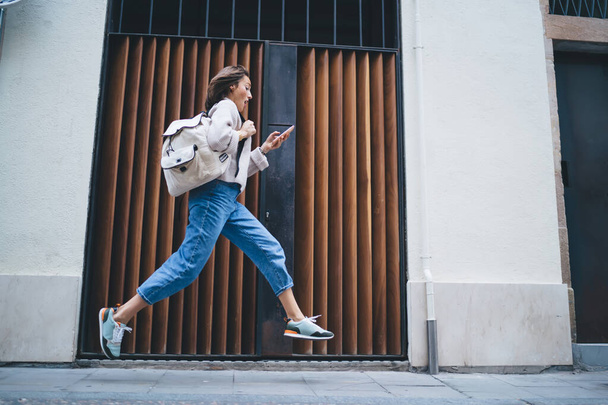 Low angle full body of anxious woman in casual clothes with backpack checking time on smartphone while running fast along street near modern building - Fotó, kép