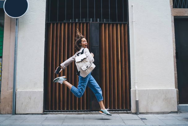 Full body of expressive middle aged female traveler in denim with backpack and smartphone in hand having fun and jumping on street during city tour - 写真・画像