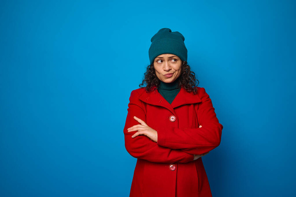 Portrait of a puzzled thoughtful pensive curly dark-haired pretty woman in red coat and green looking aside posing with crossed arms against blue wall background with copy ad space - Foto, Imagem