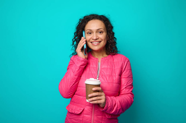 Привлекательная молодая женщина в ярко-розовой куртке держит одноразовый картонный стаканчик с горячим кофе и улыбается зубастой улыбкой, разговаривая по мобильному телефону и глядя в камеру, изолированные на синем фоне копировального пространства - Фото, изображение