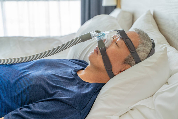 Middle age man wearing CPAP mask and headgear to help with his sleep apnea while sleeping in his bedroom - 写真・画像