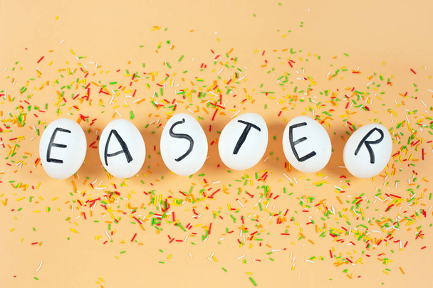 White eggs with black letters EASTER and sweet color sprinkles on a yellow pastel background. - Photo, image