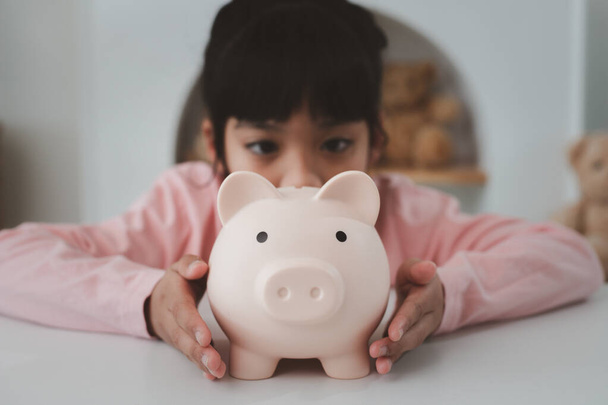 Μικρό κορίτσι από την Ασία εξοικονομεί χρήματα σε ένα κουμπαρά, μαθαίνοντας για την εξοικονόμηση, Kid εξοικονομήσει χρήματα για τη μελλοντική εκπαίδευση. Χρήματα, οικονομικά, ασφάλιση, και οι άνθρωποι έννοια - Φωτογραφία, εικόνα