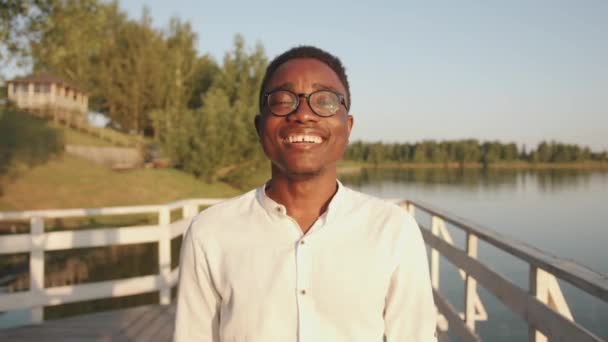 A young black guy in glasses stands on a wooden pier on the river bank and closing his eyes enjoys the sunny summer day. Slow motion - Filmati, video