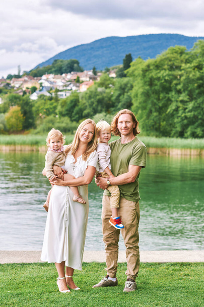 Outdoor portrait of beautiful family, young couple with preschooler boy and toddler girl posing next to lake or river - Photo, image