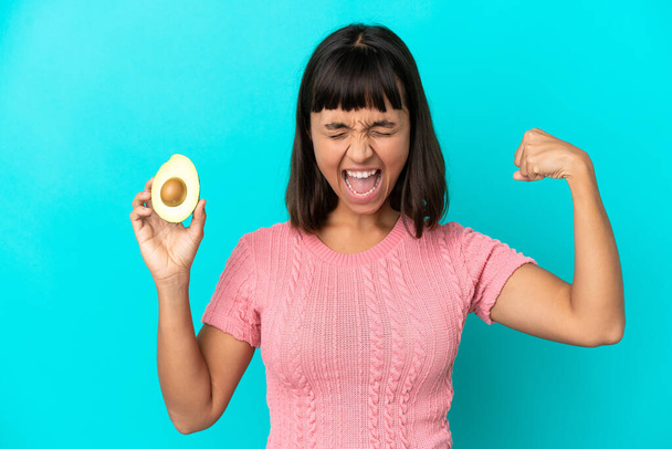 Young mixed race woman holding an avocado isolated on blue background doing strong gesture - Foto, immagini