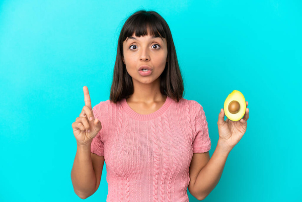 Молода змішана жінка, що тримає авокадо ізольовано на синьому фоні, має намір реалізувати рішення, піднімаючи палець вгору
 - Фото, зображення