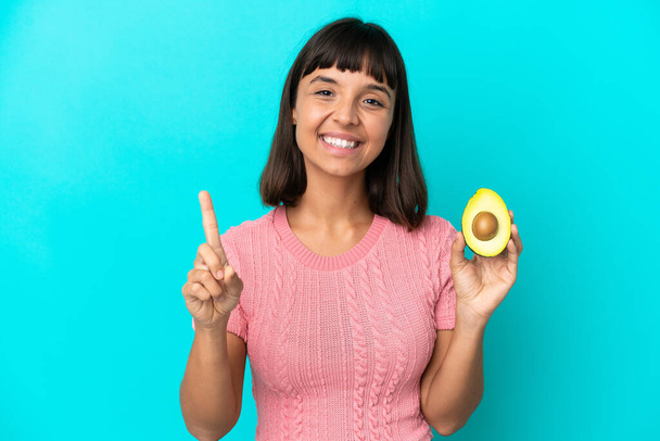 Young mixed race woman holding an avocado isolated on blue background showing and lifting a finger - Zdjęcie, obraz