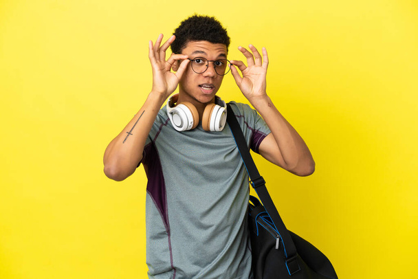 Young sport African American man with sport bag isolated on yellow background with glasses and surprised - Fotoğraf, Görsel