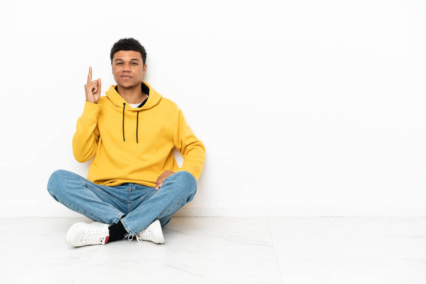 Joven hombre afroamericano sentado en el suelo aislado sobre fondo blanco señalando con el dedo índice una gran idea - Foto, Imagen