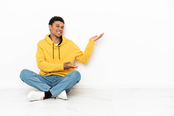 Giovane uomo afroamericano seduto sul pavimento isolato su sfondo bianco allungando le mani di lato per invitare a venire - Foto, immagini