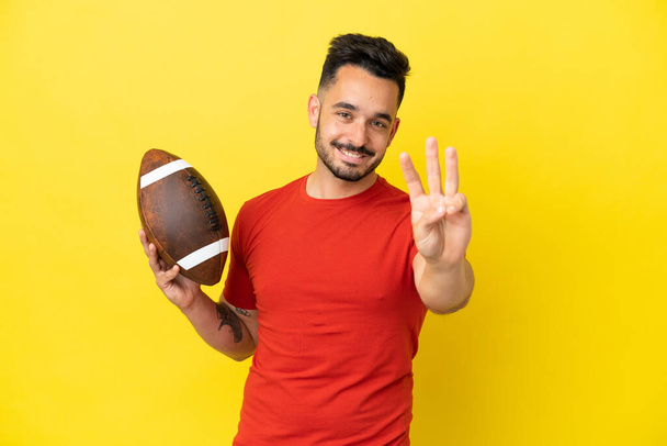 Молодой кавказский человек играет в регби на желтом фоне счастливым и считает троих с пальцами - Фото, изображение