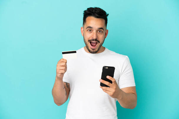 Молодой кавказский мужчина на голубом фоне покупает мобильный телефон и держит кредитную карту с удивленным выражением лица - Фото, изображение