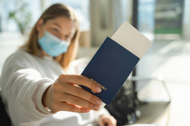 Egy maszkos fiatal nő ül egy reptéri társalgóban egy járvány idején, és bemutatja az útlevelét és a jegyét a kamerába. - Fotó, kép