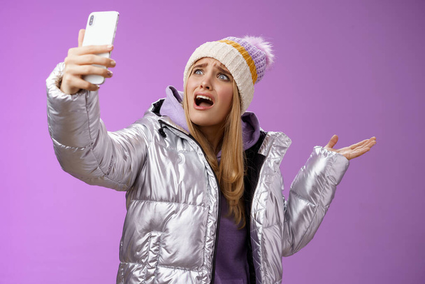 Zdenerwowana narzekająca blond dziewczyna nie może znaleźć odpowiedniego kąta robienia selfie z chłodnym zwiedzaniem podczas wakacji podróżując za granicę krzycząc smartfon wskazując na bok, fioletowe tło - Zdjęcie, obraz