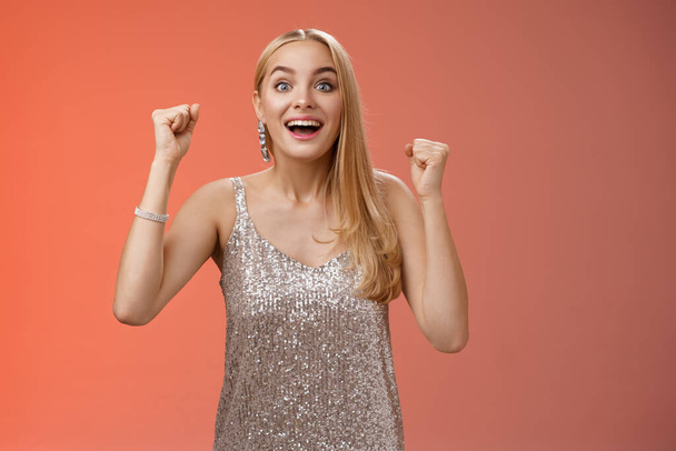 Surprised happy celebrating blond young woman in silver trendy dress raising hands up yes victory gesture smiling broadly excited winning first place reach goal grinning thrilled triumphing - Foto, Bild