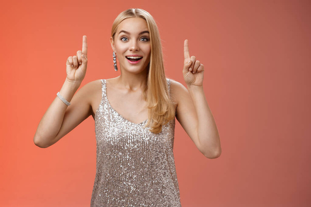 Charming amazed blond european woman in fabulous silver glittering dress raise hands point up amused enjoying watching shooting stars, fireworks gaze camera excited happy surprised, red background - Photo, image