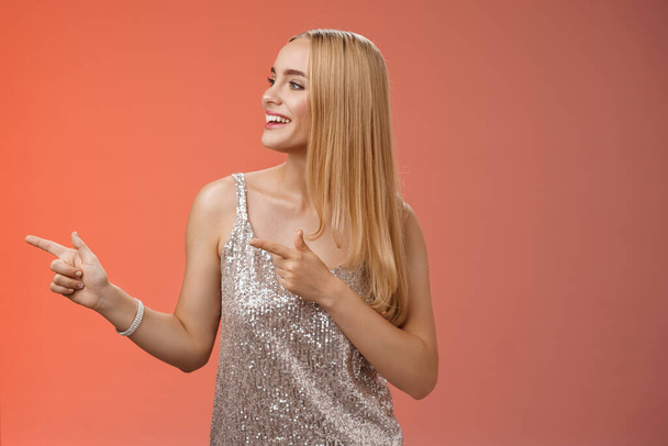 Joyful carefree young wealthy glamour blond woman in glittering silver dress enjoying awesome party dancing turning pointing left smiling broadly waiting girlfriend bring drinks, red background - Photo, Image