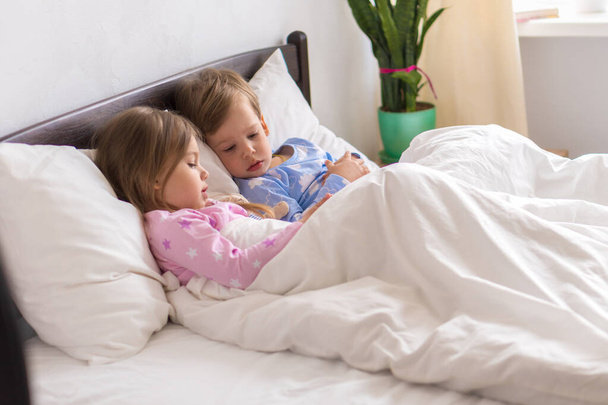 I bambini piccoli di età prescolare fratelli minori guardano insieme i cartoni animati utilizzano il dispositivo smartphone. Bambino in pigiama sul letto bianco a casa camera da letto. Famiglia, Tempo libero, Infanzia, Amicizia - Foto, immagini
