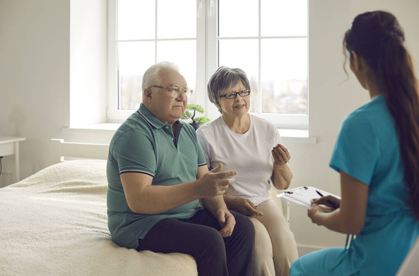 Worried senior couple sitting on bed and talking to nurse or family practitioner - Zdjęcie, obraz