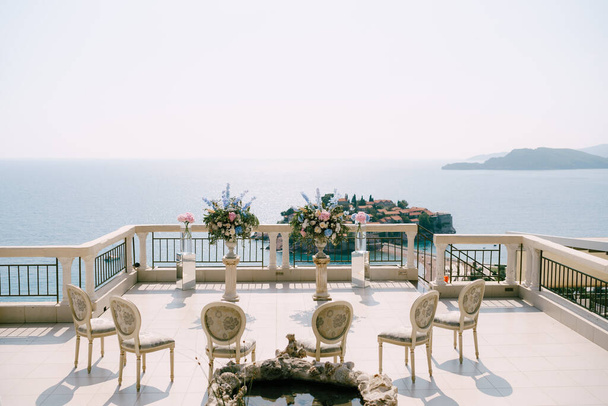 Upholstered chairs stand in front of pedestals with flowers overlooking the Sveti Stefan island  - Photo, Image