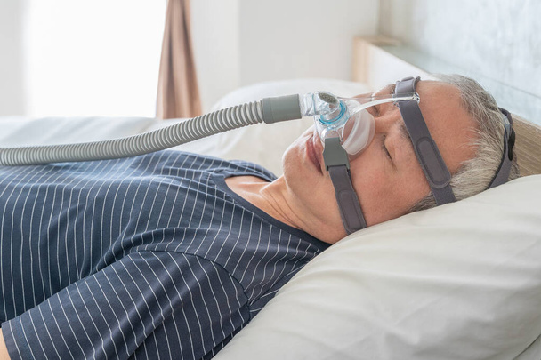 Middle age man wearing CPAP mask and headgear to help with his sleep apnea while sleeping in his bed in his bedroom - Foto, Imagen