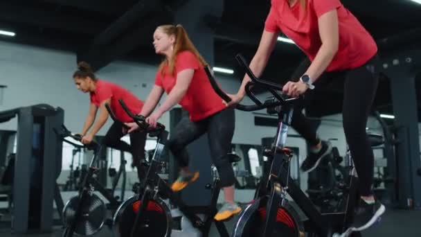 Group of smiling friends women class exercising, training, spinning on stationary bike at modern gym - Filmati, video