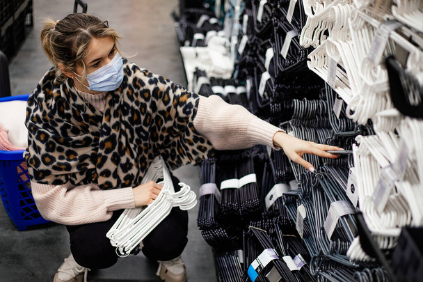 Tıbbi maskeli genç bir kadın alışveriş merkezinde soyunma odası düzenlemek için plastik askılar seçiyor. Yeni normal konsept. Salgın hastalık alışverişi. Sessizlik lütfen. Yaşam biçimi. - Fotoğraf, Görsel