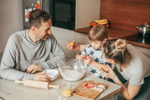 Little boy 4 - 5 years old Holds an egg in hand To break it To knead dough For baking cookies. Dad and mom are happy to watch him, real people. Family spends time together in kitchen at home. - Zdjęcie, obraz