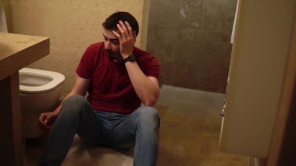 Genç sakallı sarhoş adam tuvaletin yanında oturuyor karnını tutuyor ve saf su içiyor. - Video, Çekim