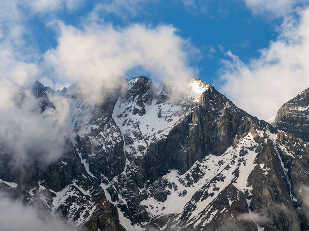 Witte wolken over een hoge puntige berg. Snowy hooggelegen alpenlandschap met besneeuwde bergtop en scherpe rotsen onder bewolkte blauwe lucht.  - Foto, afbeelding