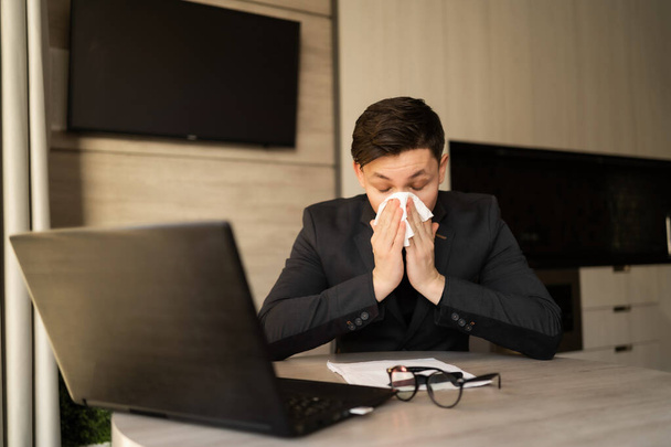 Sick young man at work got allergic to flu, sneezing and fasting, wiping a runny nose with a paper napkin, allergic guy caught a cold at work, respiratory illness sitting in home office, sick leave - Photo, Image